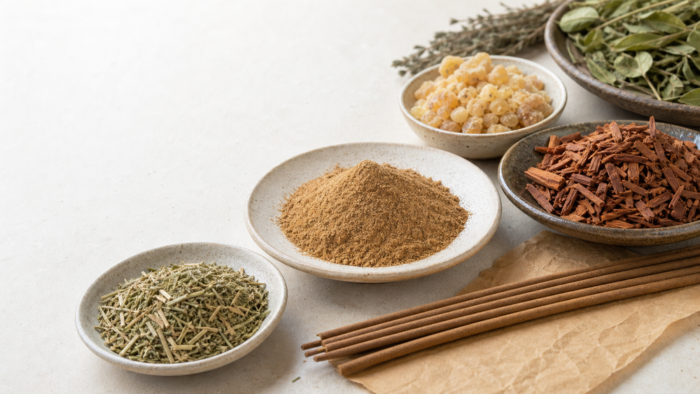 Incense sticks arranged beside sandalwood powder, wood chips, resin, and dried herbs on a light tabletop.