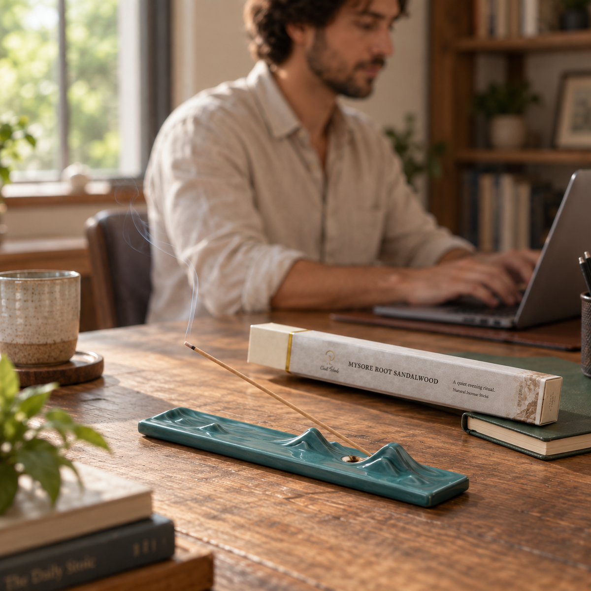Mysore Root Sandalwood incense with a dark green landscape holder on a desk, with a person working in the background